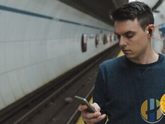 Documentary photo of a man on a subway platform using a smartphone with wired earbuds — commute mobile streaming context without Vodafone branding.