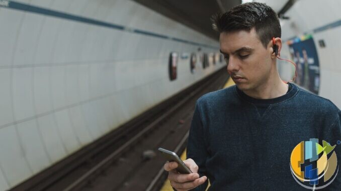 Documentary photo of a man on a subway platform using a smartphone with wired earbuds — commute mobile streaming context without Vodafone branding.