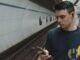 Documentary photo of a man on a subway platform using a smartphone with wired earbuds — commute mobile streaming context without Vodafone branding.