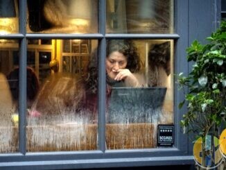 Woman working on a laptop at a table inside a café, seen through the front window