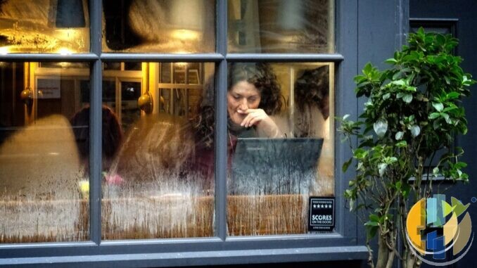 Woman working on a laptop at a table inside a café, seen through the front window
