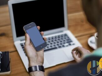 Person holding a smartphone while typing on a laptop at a wooden desk