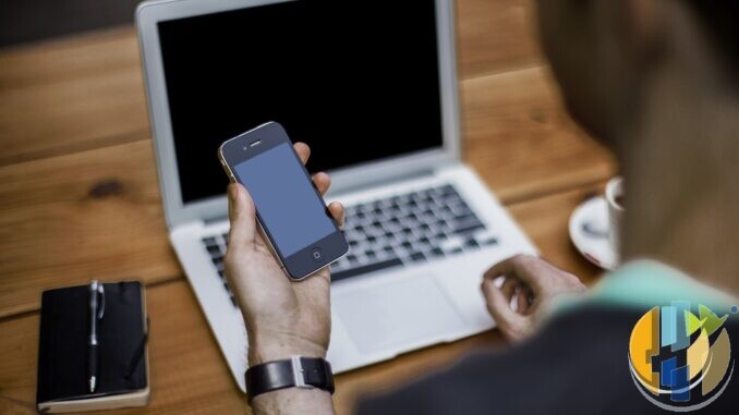 Person holding a smartphone while typing on a laptop at a wooden desk
