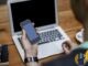 Person holding a smartphone while typing on a laptop at a wooden desk