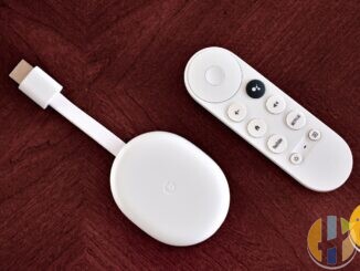 White Chromecast with Google TV dongle and its remote lying on a dark red wooden table