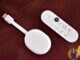 White Chromecast with Google TV dongle and its remote lying on a dark red wooden table