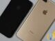 Jet black iPhone 7 and gold iPhone 7 Plus lying face down side by side on a light grey surface, showing their rear cameras and Apple logos
