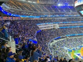 Los Angeles Rams fans in blue and yellow shirts packing the stands at SoFi Stadium during a home game, with the SoFi Stadium video board above and the field in the corner