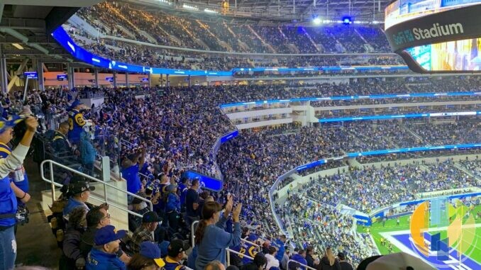 Los Angeles Rams fans in blue and yellow shirts packing the stands at SoFi Stadium during a home game, with the SoFi Stadium video board above and the field in the corner