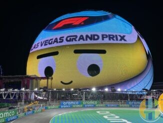 The Sphere in Las Vegas lit up at night as a giant cartoon face wearing a Las Vegas Grand Prix headband, above a floodlit corner of the street circuit lined with sponsor boards