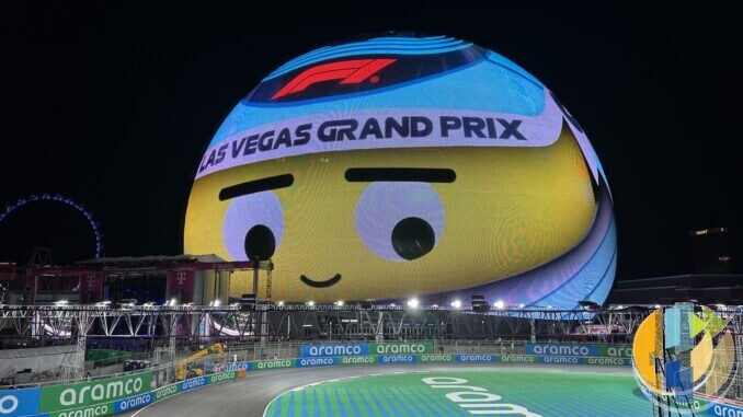 The Sphere in Las Vegas lit up at night as a giant cartoon face wearing a Las Vegas Grand Prix headband, above a floodlit corner of the street circuit lined with sponsor boards