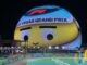 The Sphere in Las Vegas lit up at night as a giant cartoon face wearing a Las Vegas Grand Prix headband, above a floodlit corner of the street circuit lined with sponsor boards
