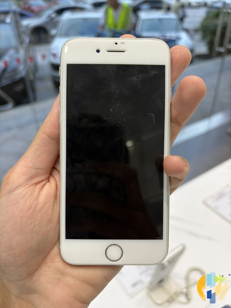 Hand holding a silver and white iPhone 6s with a dark screen and a round home button, in a shop with cars visible through the window behind